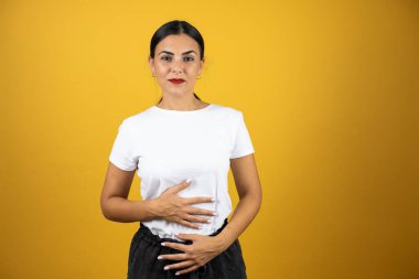 Genç ve güzel bir kadın karnına dokunuyor ve önüne bakıyor. Sarı arka planda günlük beyaz tişört ve kırmızı ruj.