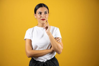 Genç bir kadın parmağını ağzının yanına koyup sağa bakarak bir şey düşünüyor. İzole edilmiş sarı bir arka planda duruyor.