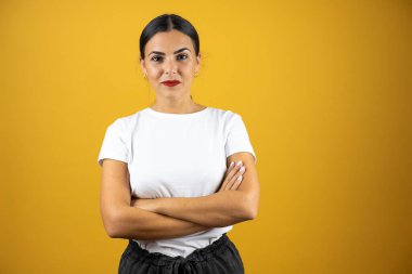 Sarı arka planda dikilmiş sıradan beyaz tişörtlü genç ve güzel bir kadın kollarını kavuşturuyor..