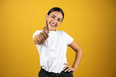 İzole edilmiş sarı arka planda genç ve güzel bir kadın bir numaralı parmağını gösterirken kendinden emin ve mutlu bir şekilde gülümsüyor.