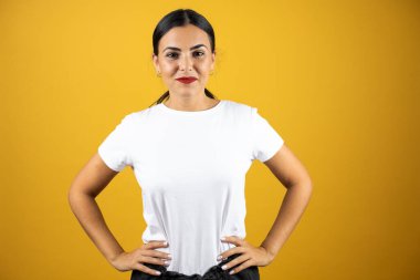 Genç ve güzel bir kadın kameraya bakıyor ve ellerini kalçalarının yanına koyuyor. Sarı arka planda günlük tişört giyiyor.