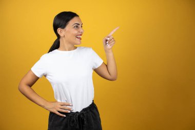 Genç, güzel bir kadın, izole edilmiş sarı arka planda başparmağıyla mutlu bir şekilde gülümsüyor.