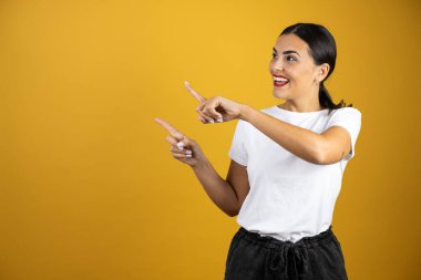 İzole edilmiş sarı arka planda rahat bir tişört giymiş güzel bir genç kadın gülümseyerek ellerini ve parmaklarını sol tarafa doğru gösteriyor.