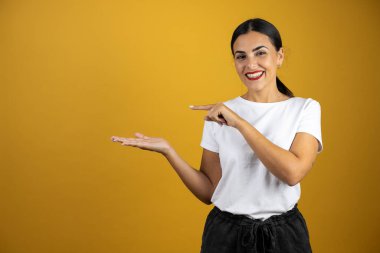 Genç ve güzel bir kadın gülümseyerek elindeki boşluğu işaret ediyor. Elindeki ürünleri gösteriyor. Sarı arka planda duruyor.