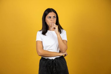 Genç, güzel bir kadın, izole edilmiş sarı arka planda duruyor. Kendine güveniyor, kollarını kavuşturup, çenesini kaldırarak düşünüyor.