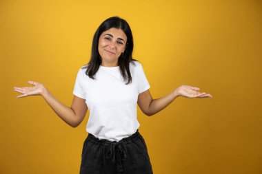 Soyutlanmış sarı arka plan üzerinde genç ve güzel bir kadın. Kolları ve elleri havada, kafası karışmış bir ifade.