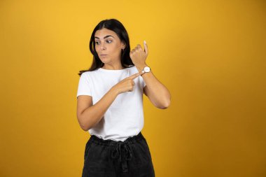 İzole edilmiş sarı arka planda duran genç ve güzel bir kadın şaşırdı ve saatini işaret etti çünkü geç oldu.