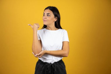 İzole edilmiş sarı arka planda duran genç ve güzel bir kadın başparmağını yana uzatıp mutlu bir şekilde gülümsüyor. Günlük beyaz tişört giyiyordu.