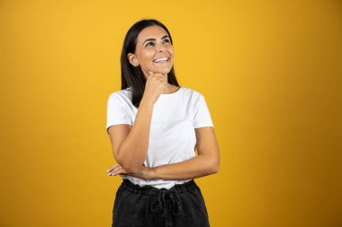 Genç, güzel bir kadın, izole edilmiş sarı arka planda duruyor. Kendine güvenen, kollarını kavuşturmuş ve çenesini kaldırmış gülümseyen bir kadın.