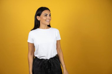 Gayri resmi beyaz tişörtlü genç ve güzel bir kadın izole edilmiş sarı arka planda dikilip sağ ön tarafa bakıyor..