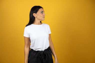 Gayri resmi beyaz tişörtlü genç ve güzel bir kadın izole edilmiş sarı arka planda dikilip sağdaki boşluğa bakıyor..