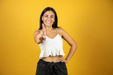 Genç, güzel bir kadın, izole edilmiş sarı arka planda duran parmaklarıyla havalı bir işaret veriyor. Beyaz günlük gömlek ve siyah şort giyiyor. Yaz görünümü