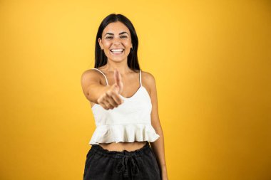 İzole edilmiş sarı arka planda duran genç ve güzel bir kadın elleriyle mutlu bir jest yapıyor. Kameraya bakıp başarıyı gösteren ifade onaylanıyor.