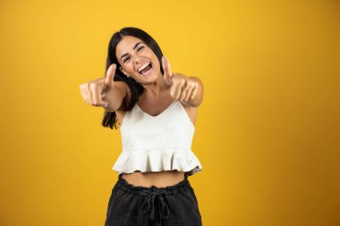 İzole edilmiş sarı arka planda duran genç ve güzel bir kadın elleriyle mutlu bir jest yapıyor. Kameraya bakıp mutluluğu gösteren yüz ifadesi.
