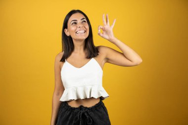 İzole edilmiş sarı arka planda duran genç ve güzel bir kadın parmaklarıyla imza atıyor ve yukarı bakıyor, mükemmel bir sembol.