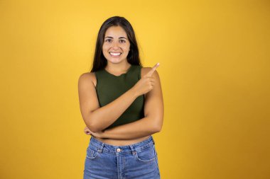 Sarı arka planda duran genç ve güzel bir kadın gülümsüyor mutlu bir şekilde elini ve parmağını yana doğru gösteriyor.