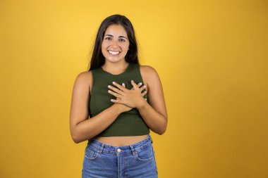 Sarı arka planda duran genç ve güzel bir kadın. Elleri göğsünde ve yüzünde minnet dolu bir jest ile gülümsüyor..