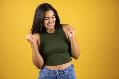 Sarı arka planda genç ve güzel bir kadın çok mutlu ve heyecanlı. Kollarını kaldırıp, gülümseyerek ve başarı için bağırarak kazanan bir jest yapıyor..