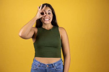 Genç, güzel bir kadın, izole edilmiş sarı arka planda dikiliyor. Gülümseyen yüzüyle, gözleriyle parmaklarının arasından bakan bir jest yapıyor.