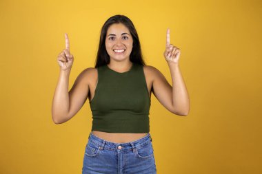 Güzel bir kadın, izole edilmiş sarı arka planda gülümsüyor, parmaklarıyla işaret ediyor ve kollarını kaldırıyordu.