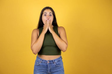Sarı arka planda duran genç ve güzel bir kadın. Elleri ağzında ve şaşırmış.