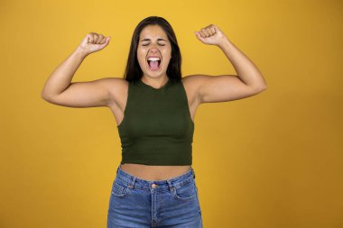 Sarı arka planda duran genç ve güzel bir kadın kolları havada bağırarak zafer işareti yapıyor.