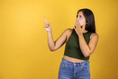 Sarı arka planda duran genç ve güzel bir kadın. Onaylanmamış bir jest yapıyor ve eli ağzında ve parmağını işaret ediyor.
