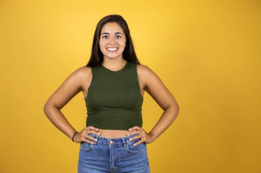 Sarı arka plan üzerinde duran genç ve güzel bir kadın. Kolları belinde gülümsüyor.