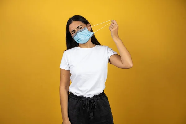 Young woman wearing the coronavirus mask. Traying to take it off because it os to tie. Standing over an isolated yellow background. Covid19