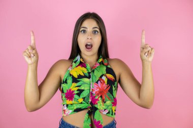 Soyutlanmış pembe arka planı olan genç ve güzel bir kadın kameraya bakıp parmaklarını kaldırıp kollarını kaldırdığında şaşırmış ve şaşırmıştı.
