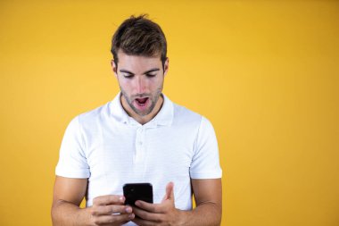 Sarı arka plan üzerinde akıllı telefon kullanan genç bir adam. Şaşırmış bir yüz, kendine güvenen bir gülümseme ve dişlerini gösteren bir gülümsemeyle.