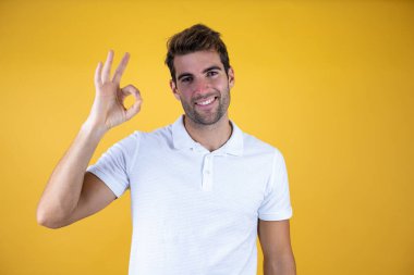 İzole edilmiş arka planda günlük beyaz tişört giyen yakışıklı genç adam gülümsüyor ve el ve parmaklarıyla işaret veriyor. Başarılı ifade