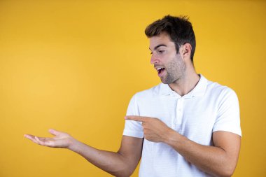 Sarı arka planda, sıradan tişörtler giyen genç yakışıklı bir adam elinde bir şeyi gösterip, parmağıyla işaret ediyor..