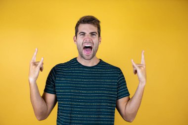 Sarı izole edilmiş arka plan üzerine gündelik tişört giyen genç yakışıklı adam elleri havada rock sembolü yaparken çılgın bir ifadeyle bağırıyor. Müzik yıldızı. Ağır konsept.