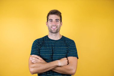 Sarı arka planda duran, kollarını kavuşturup kameraya gülümseyen, sıradan mavi tişört giyen genç yakışıklı adam. Pozitif kişi.