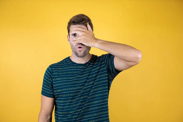 Young man standing over isolated yellow background peeking in shock covering face and eyes with hand, looking through fingers with embarrassed expression