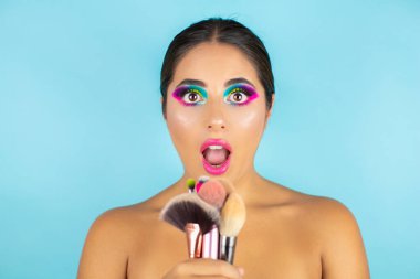 Canlı makyajlı ve mavi arka planda fırçaları olan şaşırmış bir kadının güzel portresi..