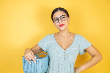 İzole edilmiş sarı arka planda duran gözlüklü güzel bir kadın çamaşır sepetini boş tutuyor.