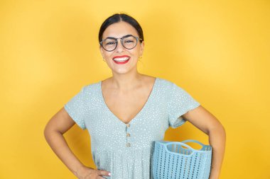 İzole edilmiş sarı arka planda duran gözlüklü güzel bir kadın çamaşır sepetini boş tutuyor.