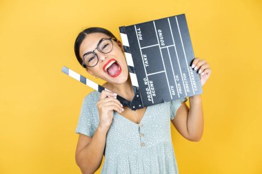 Sarı arka planda duran gözlük takan genç ve güzel bir kadın elinde klaket tutarak eğleniyor.