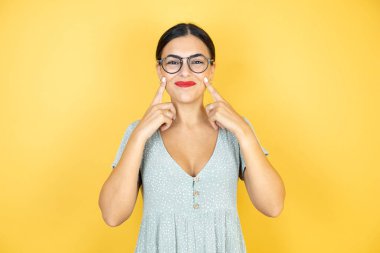 İzole edilmiş sarı arka planda gözlük takan genç ve güzel bir kadın gülümsüyor ve parmaklarıyla ve ağzıyla işaret ediyor.