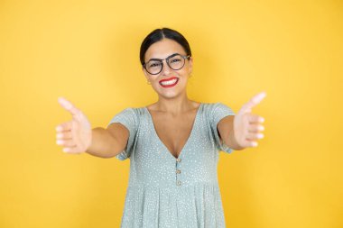 Gözlüklü güzel bir kadın, sarı arka planda, kameranın karşısında duruyor. Sarılmak için kollarını açmış gülümsüyor.