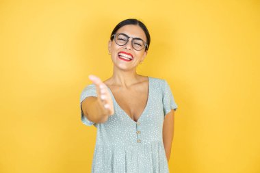 İzole edilmiş sarı arka planda duran gözlüklü genç ve güzel bir kadın dostça bir selamlaşma ve selamlama olarak el sıkışma sunuyor.