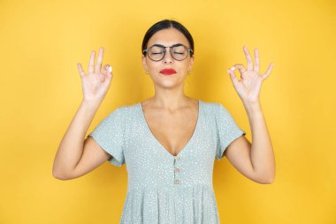 İzole edilmiş sarı arka planda duran gözlüklü güzel bir kadın rahatla ve gözün kapalı gülümse parmaklarınla meditasyon hareketi yap. Yoga konsepti.