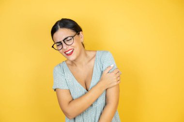 Gözlük takan genç ve güzel bir kadın izole edilmiş sarı arka planda duruyor ve acı verici omuzlara dokunuyor..