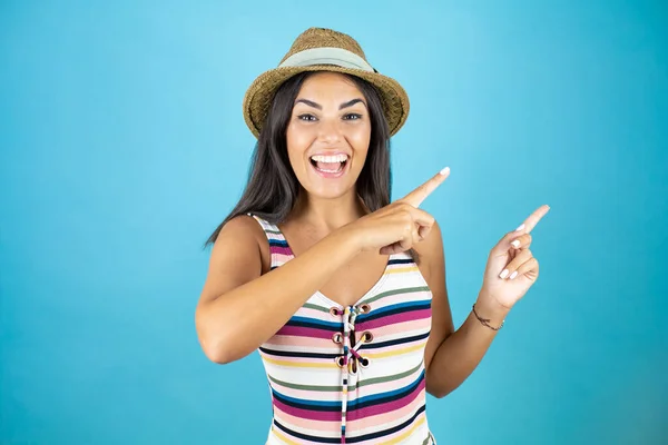 Young beautiful woman wearing swimsuit and hat over isolated blue background surprised and looking side pointing with two hands and fingers to the side.