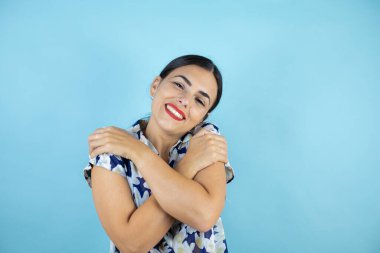Genç ve güzel bir kadın mavi arka planda, kendine sarılıyor mutlu ve pozitif, kendine güvenen..