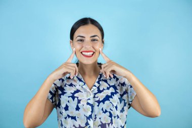 Genç ve güzel bir kadın, izole edilmiş mavi arka planda gülümsüyor ve parmaklarıyla ve ağzıyla gösterip işaret ediyor..