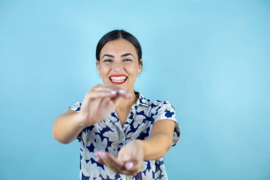 Genç ve güzel bir kadın, izole edilmiş mavi arka planda kameraya bakıyor, gülümsüyor ve büyük ve büyük boyut işareti gösteren hareketlerle işaret ediyor, ölçü sembolü..