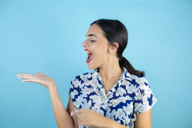 İzole edilmiş mavi arka planda duran genç ve güzel bir kadın şaşırdı ve elinde bir şey gösteriyor. Boşluğu Kopyala